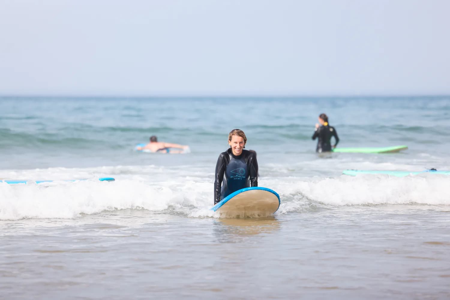 Une débutante souriante assise sur une planche bleue dans peu d’eau.