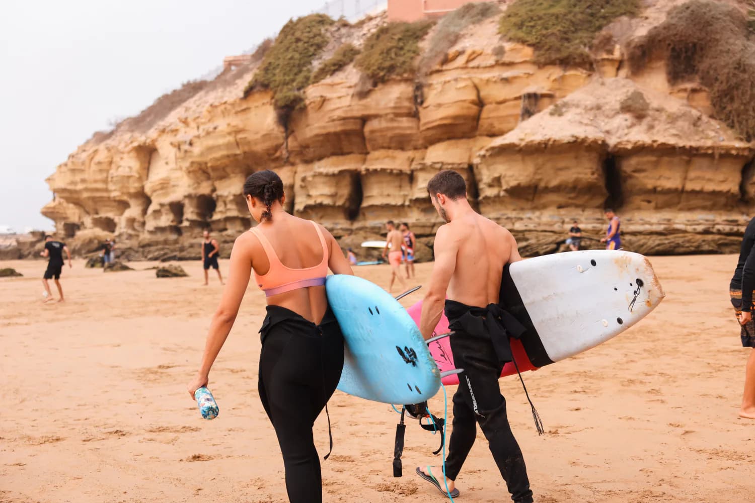Deux surfeurs portant leurs planches sous les falaises de grès stratifiées d’Imsouane.