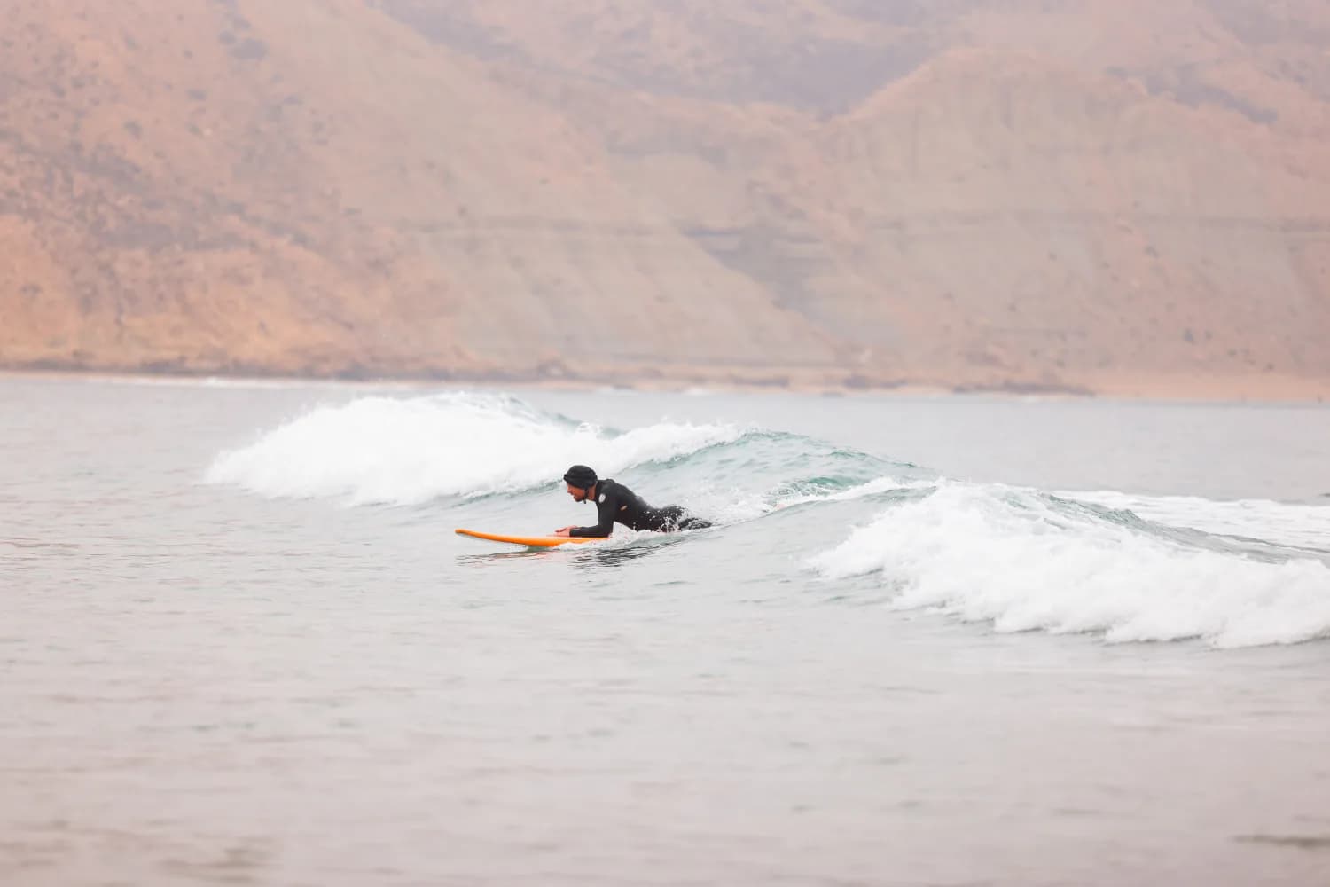 Un surfeur ramant par-dessus une petite vague avec les montagnes en arrière-plan.