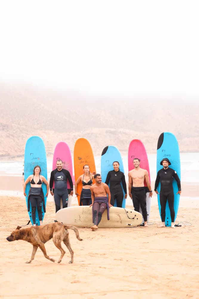 Un groupe de cours posant avec des planches colorées tandis qu’un chien traverse la plage.