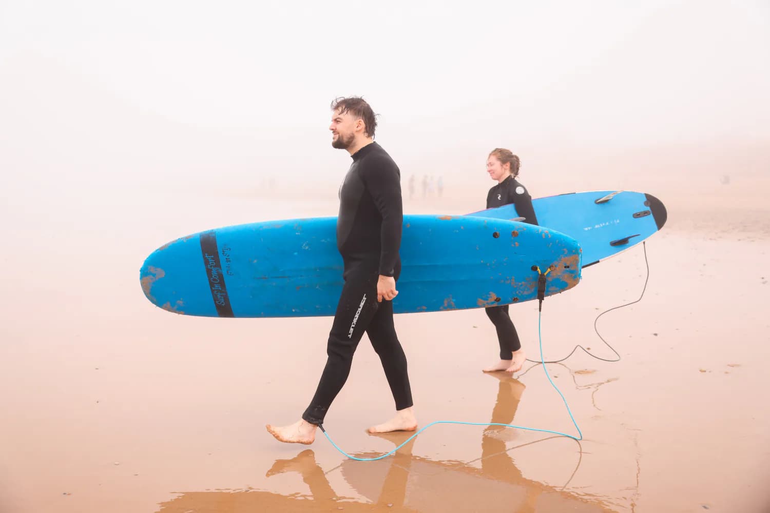 Deux surfeurs marchant sur le sable mouillé avec des planches en mousse bleues.